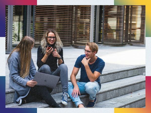 A group of people sat on steps outside with laptops, smiling and talking, framed with colourful corners.