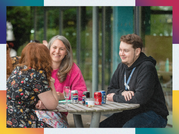 A circle of people sat at a table smiling and talking, framed with colourful corners.