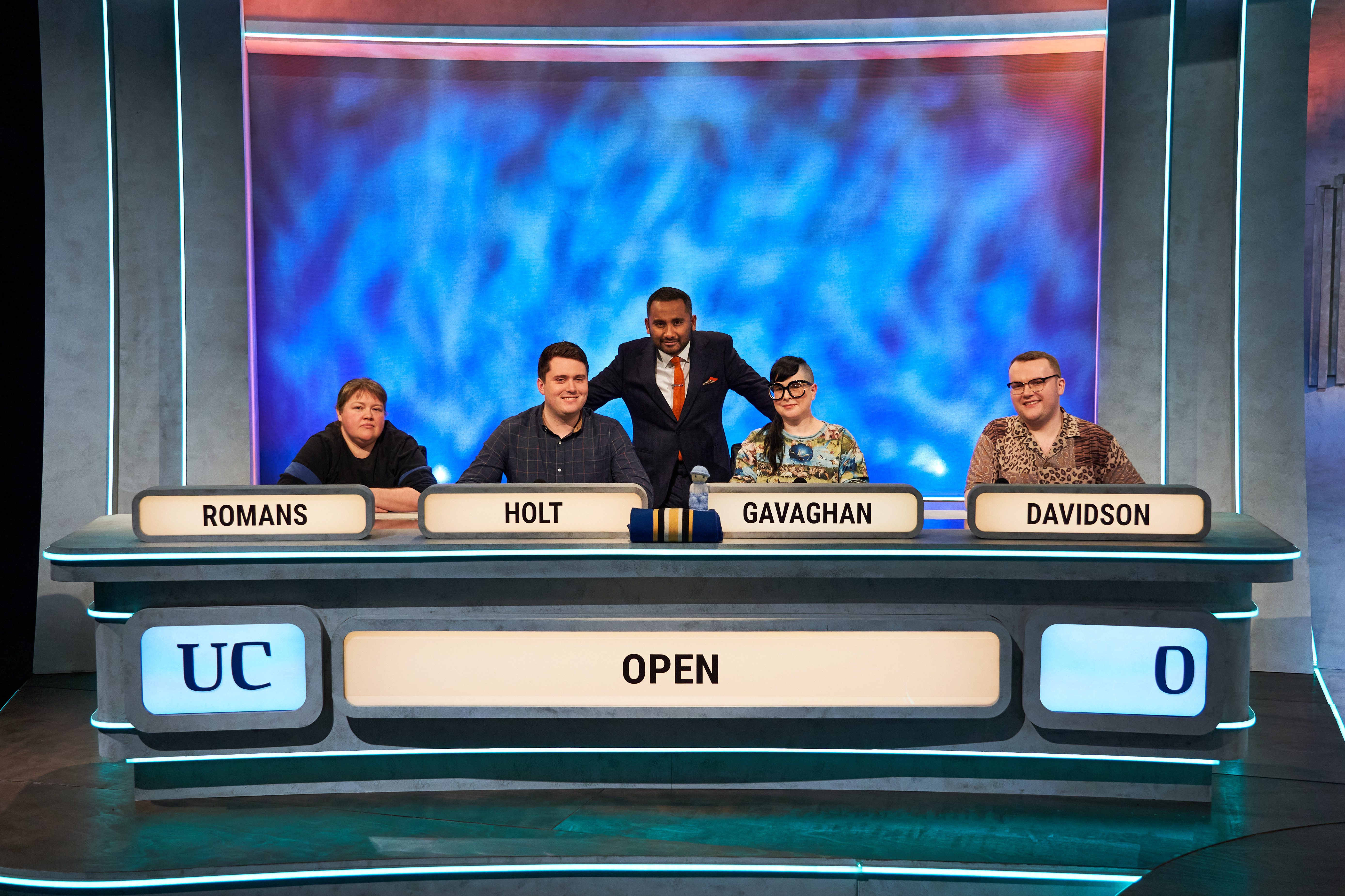 A photograph of the 2023 Open University Team on University Challenge - sat in a row at a table with their names in front of them - left to right Romans, Holt, Gavaghan and Davidson, with presenter Amol Rajan in the centre..