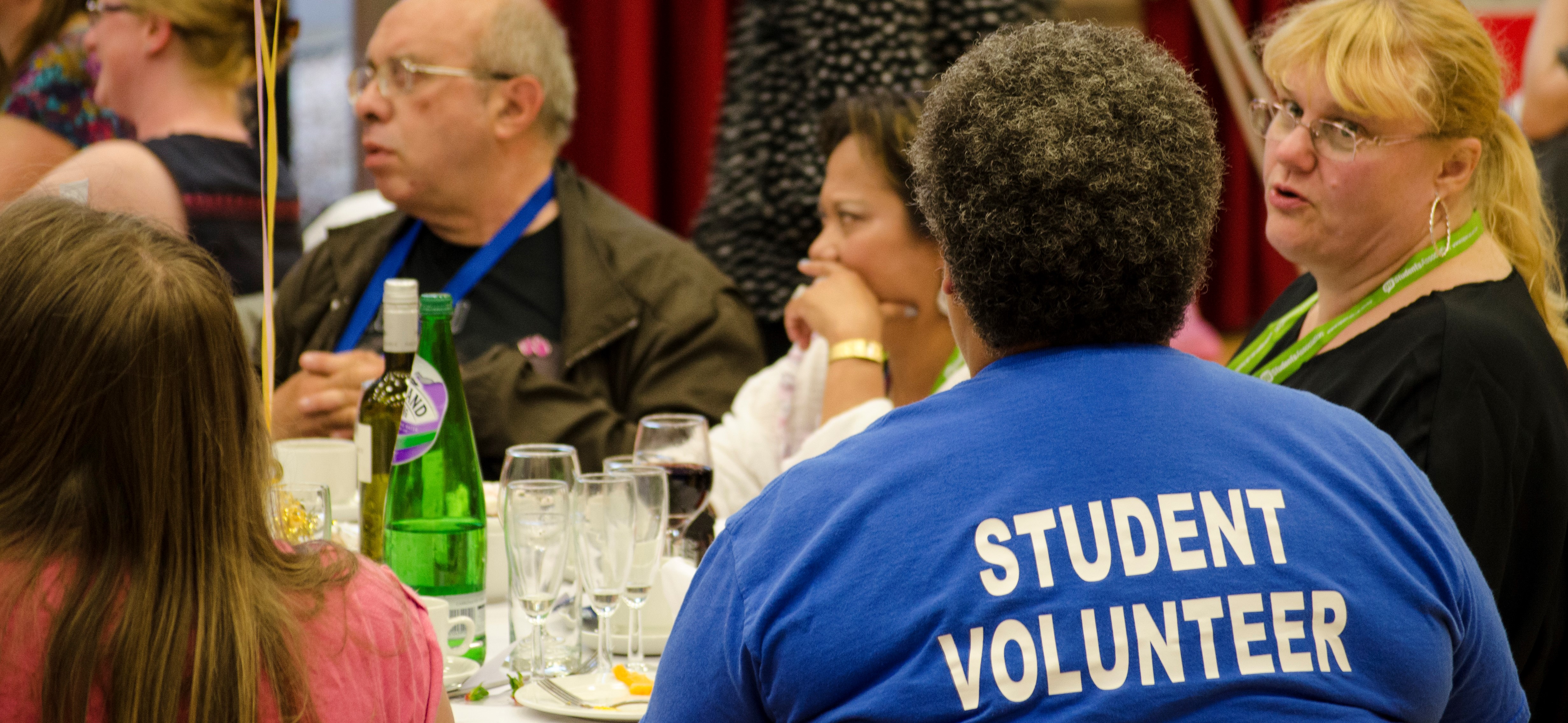 A group of 5 students sit around a table at a celebratory event. A lady with short grey and brown hair has her back to the camera in a t-shirt that reads 'student volunteer'.