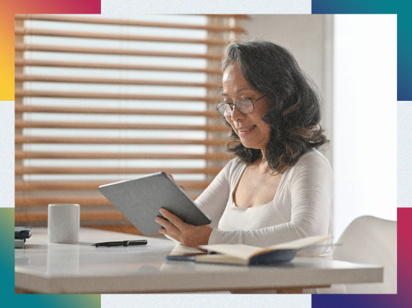 A person sat at a table smiling holding an ipad, with a notebook and mug on the table.